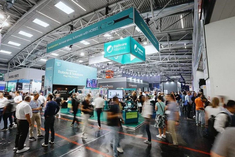 Menschenmenge auf einer Fachmesse vor dem fruitcore robotics Messestand. Im Vordergrund sind zahlreiche Besucher in Bewegung, teilweise verschwommen. Im Hintergrund der großzügig gestaltete Stand von fruitcore robotics.