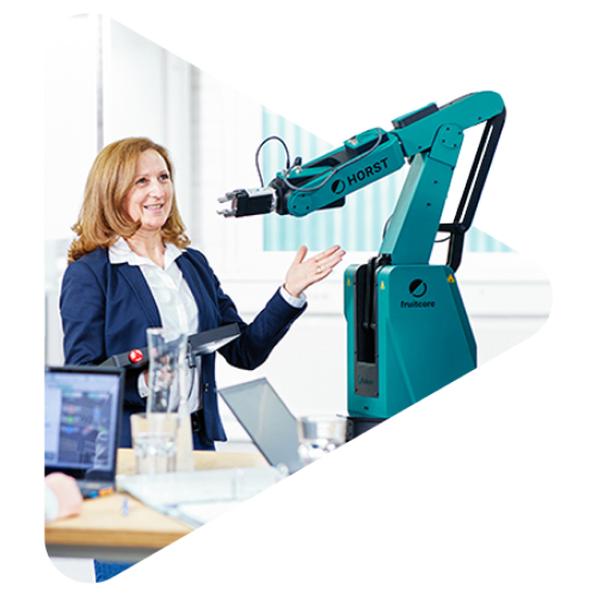 Frau in einem Büroraum hält mit dem rechten Arm ein Bedienpanel und zeigt mit der linken Hand auf einen HORST Roboter.