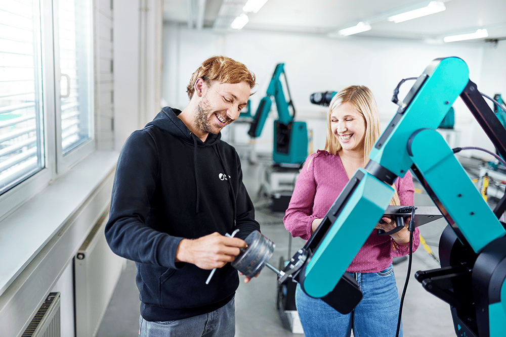 Frau und Mann stehen neben HORST Roboter und sprechen, im Hintergrund sind mehrere HORST Roboter in einer Halle zu sehen. 