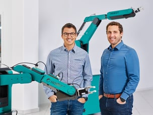 zwei Männer stehen in einem weißen Raum, im Hintergrund und im vor den Männern sind HORST Roboterarme zu sehen. 