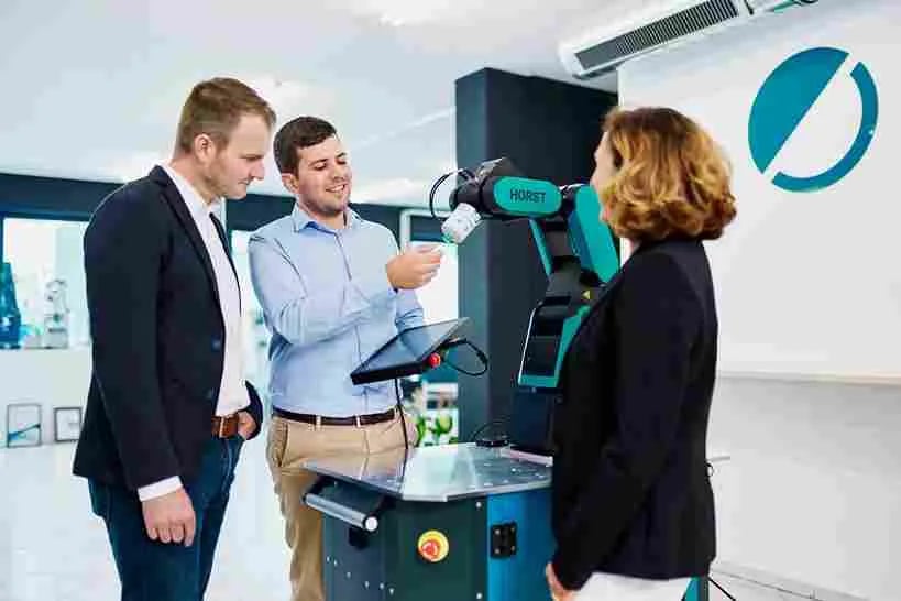 Zwei Männer und eine Frau stehen um einen HORST600 Roboter und unterhalten sich. Einer der Männer hält ein Bedienpanel in der linken Hand und zeigt mit der rechten Hand auf den Greifer des Roboters.