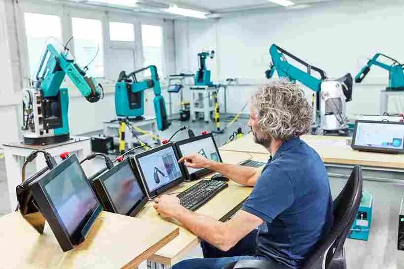 Ein Mann sitzt an einem Tisch, vor ihm liegen mehrere Bedienpanels. Er befindet sich in einer Werkstatt, in der mehrere Roboter stehen.