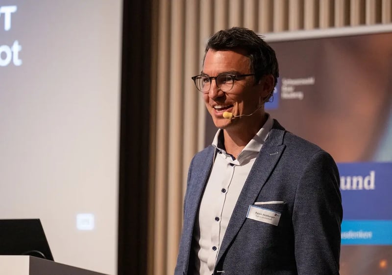 Ein Mann in Businesskleidung steht lächelnd auf einer Bühne und hält einen Vortrag. Er trägt ein Headset mit Mikrofon, eine Brille und ein Namensschild. Im Hintergrund sind ein Präsentationsbildschirm sowie ein Roll-Up mit Schriftzug erkennbar.