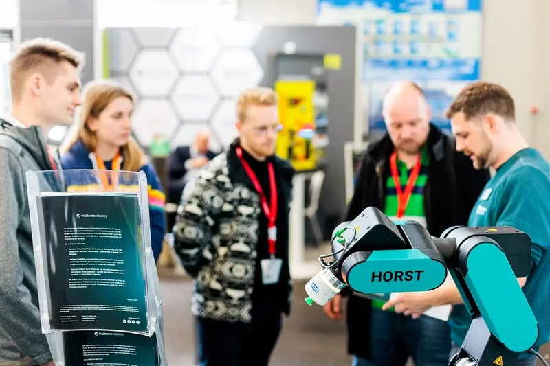 Messestand von fruitcore robotics mit einem HORST-Industrieroboter im Vordergrund, während ein Mitarbeiter einer Gruppe interessierter Besucher die Technologie demonstriert. 
