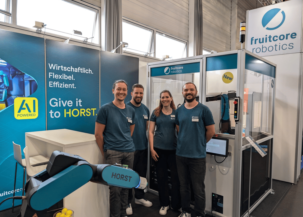 Vier Personen stehen in einem Messestand. Vor den Personen ich HORST Roboterarm zu sehen. Hinter den Personen ist eine Automatisierungslösung zu sehen in der ein HORST1500 Roboter steht. 